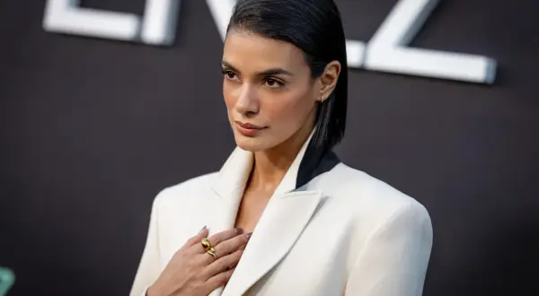 Laysla de Oliveira, elegant in a white blazer, poses with a hand on her chest, adorned with a striking gold ring.