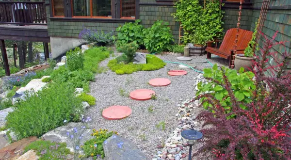 Inexpensive stepping stone walkway ideas: red circular pavers create a path through a gravel garden with lush greenery and a wooden chair.