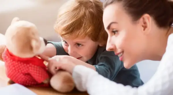 Uśmiechnięta opiekunka bawi się z dzieckiem pluszowym misiem. Zastanawiasz się, ile bierze opiekunka do dzieci za taką troskę?