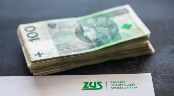 Stos banknotów 100 zł obok dokumentu ZUS. Może to symbolizować zarabianie na kontach bankowych lub oszczędzanie na przyszłość.