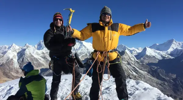 Dwóch alpinistów na szczycie Mera Peak, jeden z czekanem, drugi z kciukiem w górę. W tle ośnieżone góry.