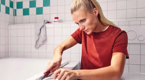 Frau entfernt mit einem Cuttermesser alte Silikonfugen an der Badewanne.