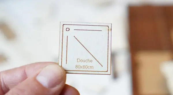 Main tenue par un pouce, une petite carte en bois gravée indique "Douche 80x80cm", une dimension pour une future douche.