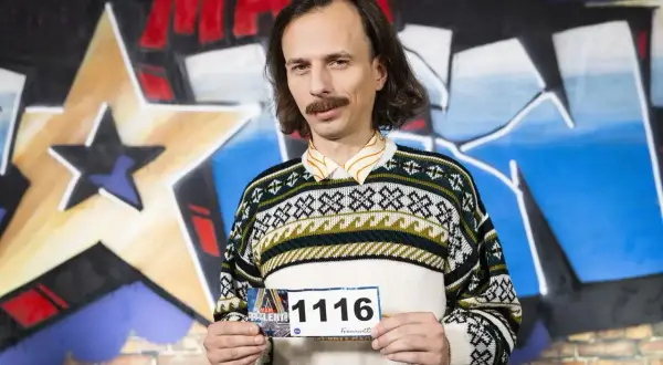 Mężczyzna z wąsami i w swetrze z geometrycznym wzorem trzyma tabliczkę z numerem 1116. Savyan czeka na swój występ.