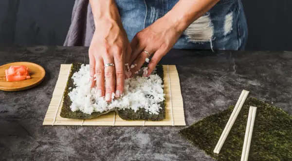 Dłonie rozprowadzają ryż do sushi na nori. Obok leżą pałeczki i marynowany imbir.