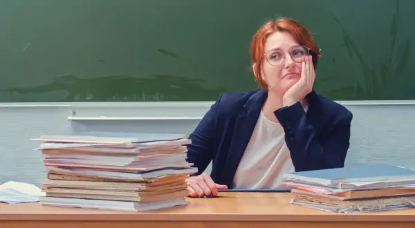 Nauczycielka zamyślona przy biurku, stosy papierów obok. Czy doczekamy się podwyżki dla nauczycieli? Najnowsze wiadomości.