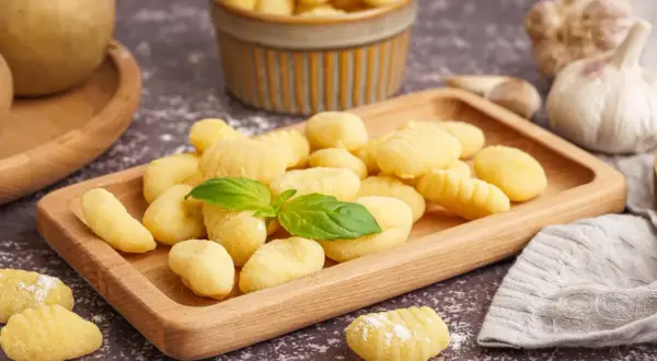 Domowe gnocchi, idealne do przygotowania według prostego przepisu. Na drewnianej desce świeże kluseczki ziemniaczane, ozdobione listkiem bazylii.