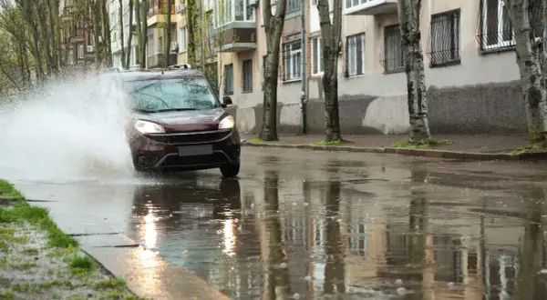 Czerwony samochód wpada w kałużę, tworząc fontannę wody. To przykład aquaplaningu, czyli utraty przyczepności na mokrej nawierzchni.