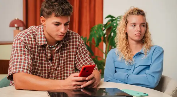 Een jongeman kijkt geconcentreerd naar zijn telefoon, terwijl zijn partner met gekruiste armen en een norse blik wegkijkt. Het voelt alsof hij haar compleet negeert, alsof ze leeggezogen wordt door zijn onoplettendheid.