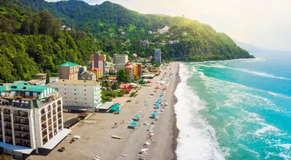 Widok na wybrzeże Gruzji, gdzie zielone wzgórza schodzą do piaszczystej plaży i turkusowego morza. Widać hotele i turystów odpoczywających nad wodą.