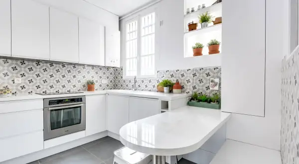 Cuisine moderne aux meubles blancs épurés, rehaussée par une crédence aux motifs géométriques gris et blancs. Une table blanche intégrée et des plantes apportent une touche de fraîcheur.