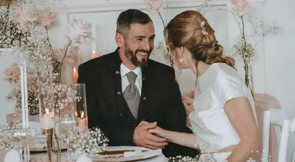 Ein glückliches Brautpaar bei ihrer Petersilienhochzeit, umgeben von zarten rosa Rosen und Schleierkraut. Sie halten Händchen und blicken sich liebevoll an.