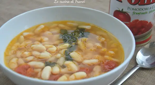 Zuppa di fagioli spollichini fumante in una ciotola bianca, con pomodori e verdure. Accanto, una lattina di pomodori e un cucchiaio.