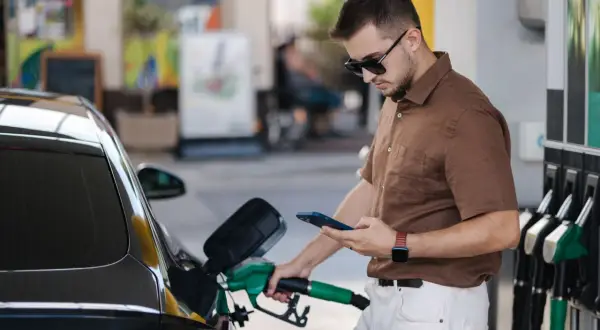 Un homme fait le plein de sa voiture à une **téléphone station essence**, consultant son smartphone.