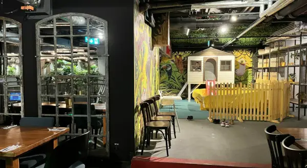Warszawska restauracja z salą zabaw dla dzieci. Widać stoliki, krzesła i kolorowy plac zabaw z domkiem i zjeżdżalnią.