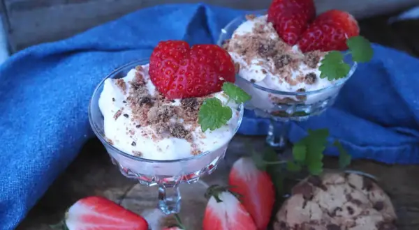 Två glas med krämig dessert toppad med jordgubbar och smulor. Bredvid ligger halverade jordgubbar och chokladkakor. En härlig sommardessert med jordgubbar och glass.