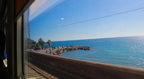 Vy från tågfönstret under en resa för att tågluffa i Italien. Blå himmel, glittrande hav och en stenstrand med palmer.
