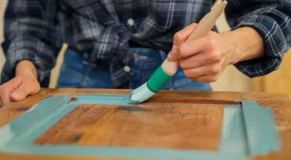 Person streicht mit Pinsel und hellblauer Farbe über Holz. Ein DIY-Projekt, um alten Möbeln neuen Glanz zu verleihen. Lack auf Lack streichen.
