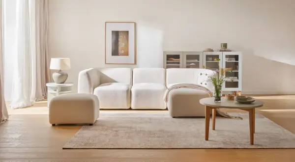 Przytulny salon z kanapą i pufą w odcieniach beżu. Beżowy kolor ścian tworzy ciepłą atmosferę.