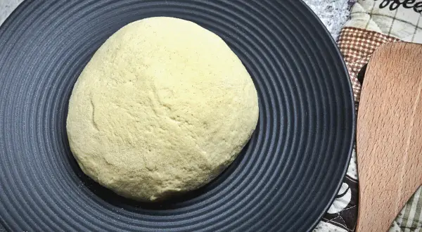 Ein runder, heller pizzateig wie beim Italiener liegt auf einem schwarzen Teller. Daneben ein Holzspatel und eine Kaffeetasse-Serviette.