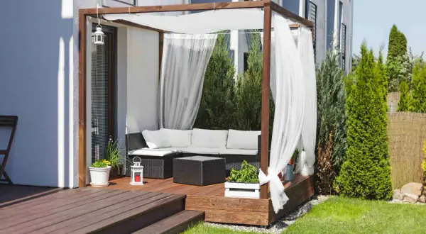 Drewniana pergola z białymi zasłonami to idealne miejsce na tarasie. Zapewnia cień i prywatność, a także stanowi stylowe tło dla mebli ogrodowych.