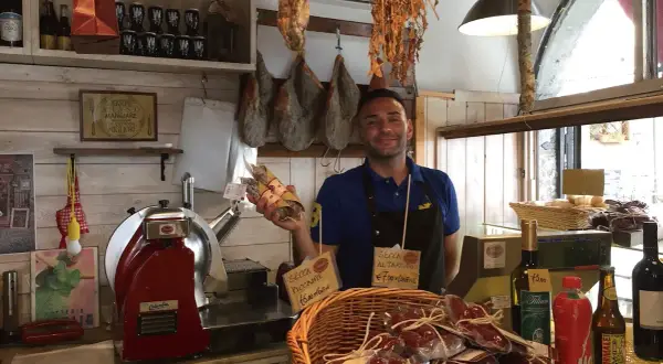 Un salumiere sorridente mostra salumi in un negozio di Bolsena. Prosciutto appeso, affettatrice e prodotti tipici invitano a scoprire i sapori locali.