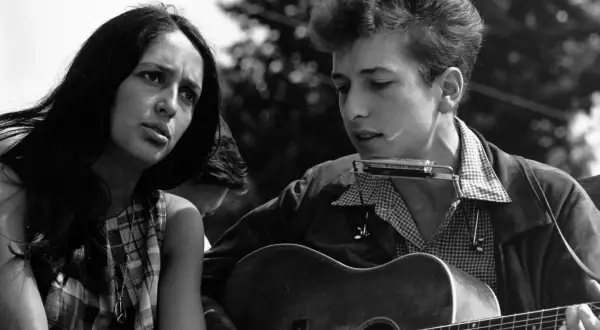 Młody Bob Dylan gra na gitarze i harmonijce, obok siedzi kobieta. To obraz z czasów, gdy jego twórczość zasłużyła na nagrodę Nobla.