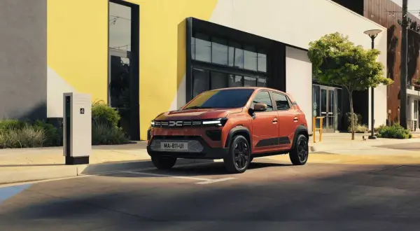En orange Dacia står parkerad vid en laddstation framför en modern byggnad. Är Dacia bra eller dålig? Denna ser lovande ut!