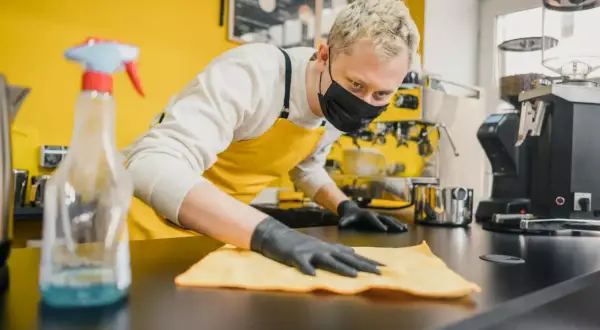 Barista con mascherina e guanti effettua la pulizia della cucina del ristorante, igienizzando il bancone con un panno giallo.
