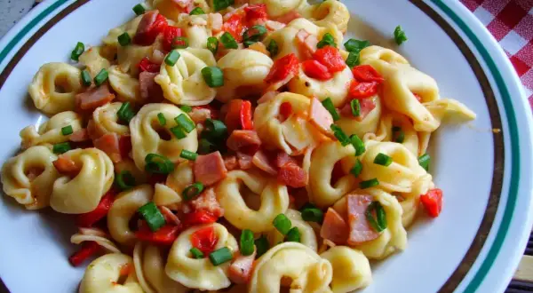 Pyszne tortellini co to jest? Makaron z kawałkami szynki, papryki i szczypiorku na talerzu.