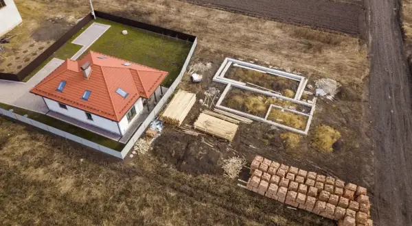 Zabudowa zagrodowa w budowie: gotowy dom z czerwonym dachem, fundamenty nowego budynku, drewno i palety cegieł.