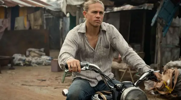 Charlie Hunnam kör motorcykel genom ett slumområde, med en blick av beslutsamhet.