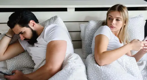 Ein Mann liegt im Bett und schaut auf sein Handy, während seine Frau ihn misstrauisch beobachtet. Er guckt vielleicht gerade Porno.