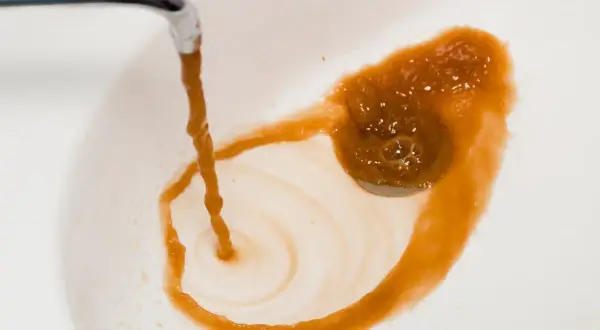 Eau brune et trouble coule d'un robinet dans un lavabo blanc, laissant des traces rouille. On dirait du poison dans l'eau.