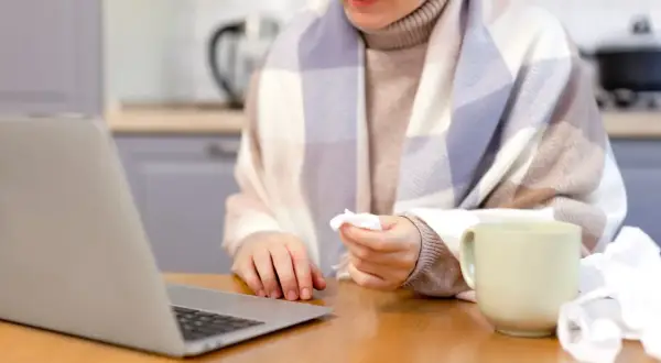 Osoba owinięta kocem, z chusteczką w dłoni, pracuje przy laptopie. Obok kubek i zużyte chusteczki. Może to być sytuacja związana z art. 148 kp.