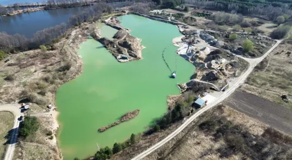 Widok z lotu ptaka na turkusowe jezioro zajączkowskie, otoczone piaskowymi skarpami i zielenią. Widać sprzęt wydobywczy i łódź.