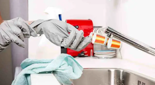 Mani con guanti grigi si preparano a sturare il lavandino, con un panno azzurro e una macchinetta del caffè rossa sullo sfondo.