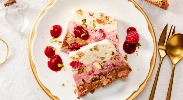Gefrorener Kuchen mit Beeren, Nüssen und Schokolade auf einem Teller mit Sauce und Löffel.