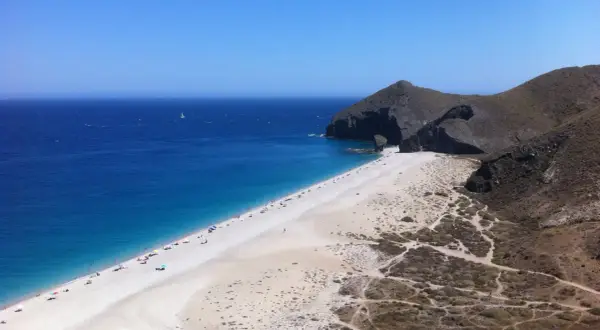 Playa de arena blanca y mar azul intenso, con acantilados rocosos y vegetación árida. Un velero navega en la distancia, un capricho del paisaje.