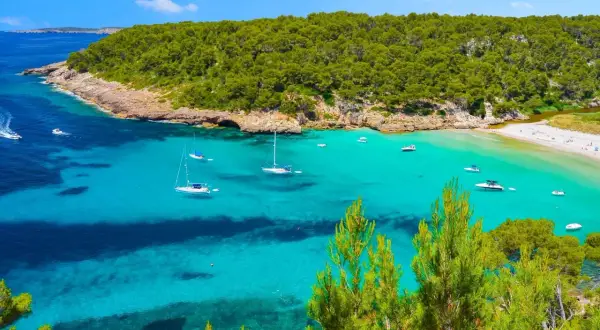 Bahía de aguas turquesas y cristalinas, con veleros y lanchas navegando. Al fondo, una playa de arena blanca y una frondosa vegetación que llega hasta la cala pudent.