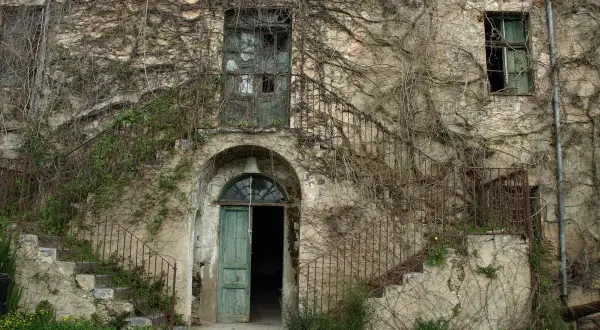 Antica casa in Sardegna, ricoperta di edera, con scale esterne arrugginite e porta verde aperta. Un sogno per chi cerca case a 1 euro.