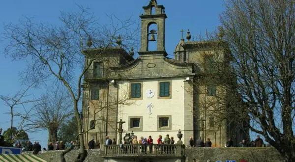 Edificio religioso con campanario y cruz. Gente en el balcón y en la plaza. Un árbol desnudo en primer plano.