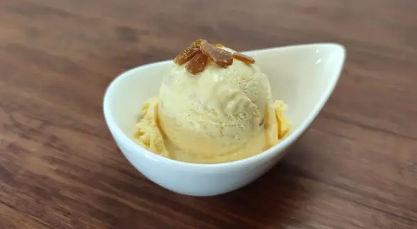 Cremiges Eis mit Eigelb, garniert mit karamellisierten Stückchen, in einer weißen Schale auf Holzhintergrund.