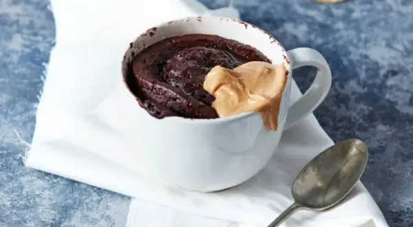 Schokoladiger high protein tassenkuchen mit Erdnussbutter-Topping in einer weißen Tasse, daneben ein Löffel.