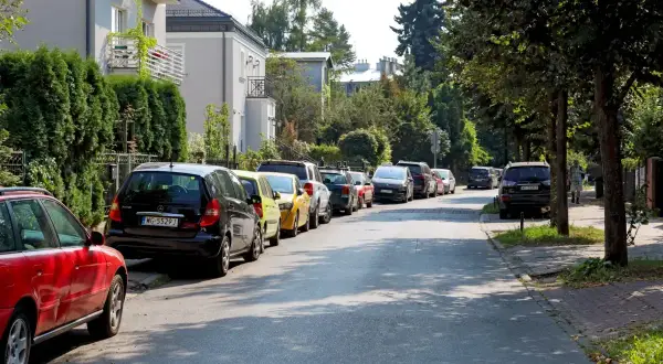 Ulica z wieloma samochodami, gdzie parkowanie na chodniku jest powszechne. Widać domy i drzewa.