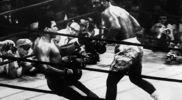 Joe Frazier fights in the ring, delivering a powerful blow as his opponent stumbles. The crowd watches intently.