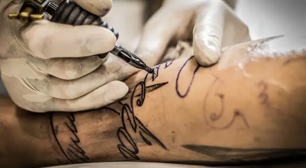 Manos enguantadas trabajan con una máquina de tatuar sobre un brazo, delineando un diseño. Aprende como cuidar un tatuaje recien hecho.