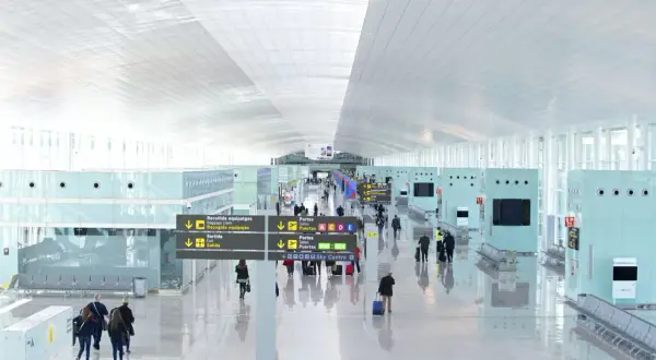 Jasny, nowoczesny terminal lotniska w Hiszpanii. Pasażerowie z bagażami przemieszczają się pod wielkim, białym sufitem, kierując się znakami.
