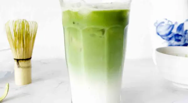 En snygg, isig matcha latte med lager av mjölk och grönt te. Bredvid står en visp för att göra matcha.