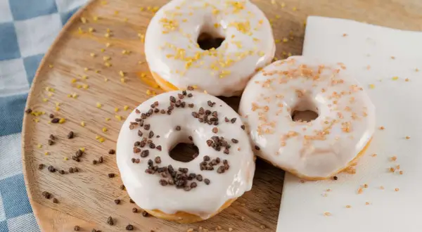 Drei Donuts mit weißer Glasur und Streuseln aus Schokolade, Zucker und Zitrone liegen auf einem Holzbrett.